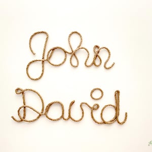 May include: The names John and David are written in cursive script using a single length of brown twisted rope. The letters are formed by the rope, creating a textured, three-dimensional appearance against a plain, light-colored background.
