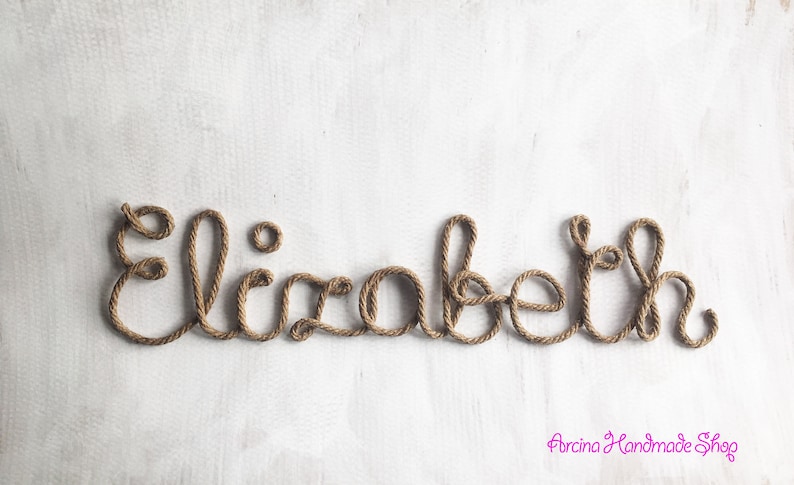 May include: The name Elizabeth is written in cursive script using a single continuous length of brown twisted rope. The letters are formed by the rope, creating a decorative wall sign. The text Arcina Handmade Shop appears in pink in the corner.