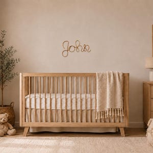 May include: A light wood crib with vertical slats, a white mattress, and a cream-colored fringed throw blanket. Mounted on the wall above the crib is a decorative wire script sign that spells out the name John in a cursive, looping font style.