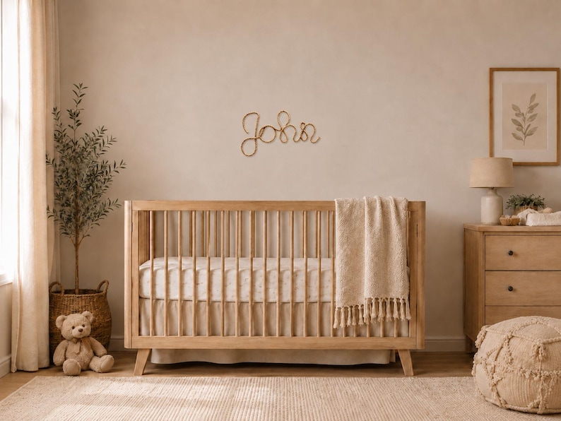 May include: A light wood crib with vertical slats, a white mattress, and a cream-colored fringed throw blanket. Mounted on the wall above the crib is a decorative wire script sign that spells out the name John in a cursive, looping font style.