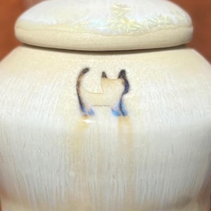 May include: A cream-colored ceramic jar with a matching lid. The jar features a hand-painted cat design in blue and black. The jar has a rounded shape and a smooth, glossy finish. The lid is oval and sits on top of the jar.
