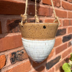 May include: A conical ceramic hanging planter featuring a speckled tan upper section and a smooth white glazed lower section. The planter is suspended by three strands of natural jute twine threaded through holes near the top rim of the vessel.
