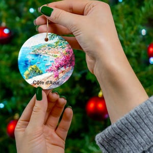 Peut inclure: Une d&eacute;coration de No&euml;l ronde en c&eacute;ramique orn&eacute;e d'une peinture aquarelle color&eacute;e repr&eacute;sentant une ville c&ocirc;ti&egrave;re m&eacute;diterran&eacute;enne avec de l'eau bleue, des voiliers et des fleurs roses. Le texte C&ocirc;te d'Azur est imprim&eacute; en noir en bas.