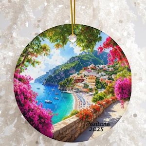 Pode incluir: Enfeite de Natal redondo em cerâmica com uma vista panorâmica de Positano, Itália, com uma cidade costeira, água turquesa e flores cor-de-rosa. O enfeite tem um cordão dourado para pendurar e o texto "Positano 2025".