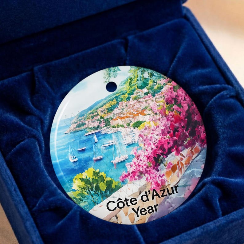 Peut inclure: Un ornement en c&eacute;ramique rond pr&eacute;sentant une peinture c&ocirc;ti&egrave;re color&eacute;e de la C&ocirc;te d'Azur avec des voiliers sur l'eau bleue, des fleurs roses et des collines vertes. Le texte C&ocirc;te d'Azur Year est imprim&eacute; en noir au bas de la surface circulaire.