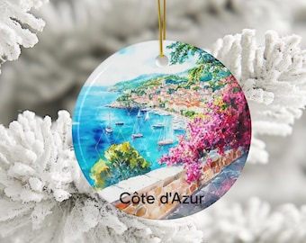 May include: A round ceramic ornament featuring a vibrant watercolour painting of the Côte d'Azur coastline, including blue water, sailboats, a hillside town, and bright pink bougainvillea flowers. The text Côte d'Azur is printed at the bottom.