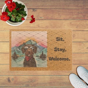Può includere: Un zerbino rettangolare in fibra di cocco con un cane con occhiali da sole e bandana rossa, sullo sfondo di una montagna e di un lago. Il testo "Sit. Stay. Welcome." è stampato. Lo zerbino è su una superficie di legno.