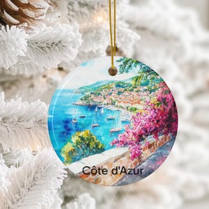 Peut inclure: Une d&eacute;coration en c&eacute;ramique ronde illustrant un paysage c&ocirc;tier de la C&ocirc;te d'Azur avec des eaux bleues, des voiliers, un village &agrave; flanc de colline et des fleurs de bougainvilliers rose vif, suspendue par un fin cordon dor&eacute;.