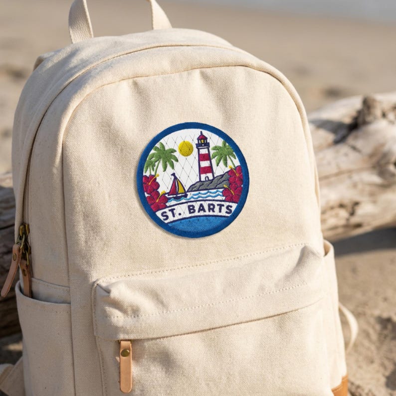 May include: A beige canvas backpack featuring a circular embroidered patch on the front pocket. The patch depicts a red and white lighthouse, a sailboat, palm trees, and red hibiscus flowers with the text ST. BARTS printed in blue letters.