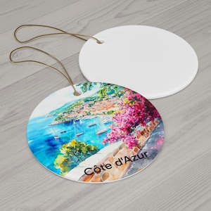 Peut inclure: Deux ornements ronds en c&eacute;ramique avec des cordons dor&eacute;s. L'un est blanc uni, tandis que l'autre pr&eacute;sente une peinture aquarelle color&eacute;e d'une ville c&ocirc;ti&egrave;re avec de l'eau bleue, des bateaux, des fleurs roses et le texte C&ocirc;te d'Azur imprim&eacute;.
