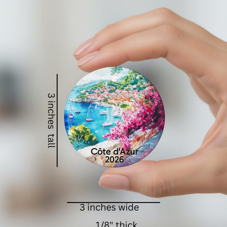 Peut inclure: Une d&eacute;coration ronde en c&eacute;ramique illustrant une peinture c&ocirc;ti&egrave;re de la C&ocirc;te d'Azur avec des voiliers et des fleurs roses. Le texte C&ocirc;te d'Azur 2026 est imprim&eacute; en bas. Elle mesure 7,62 cm de haut, 7,62 cm de large et 0,32 cm d'&eacute;paisseur.