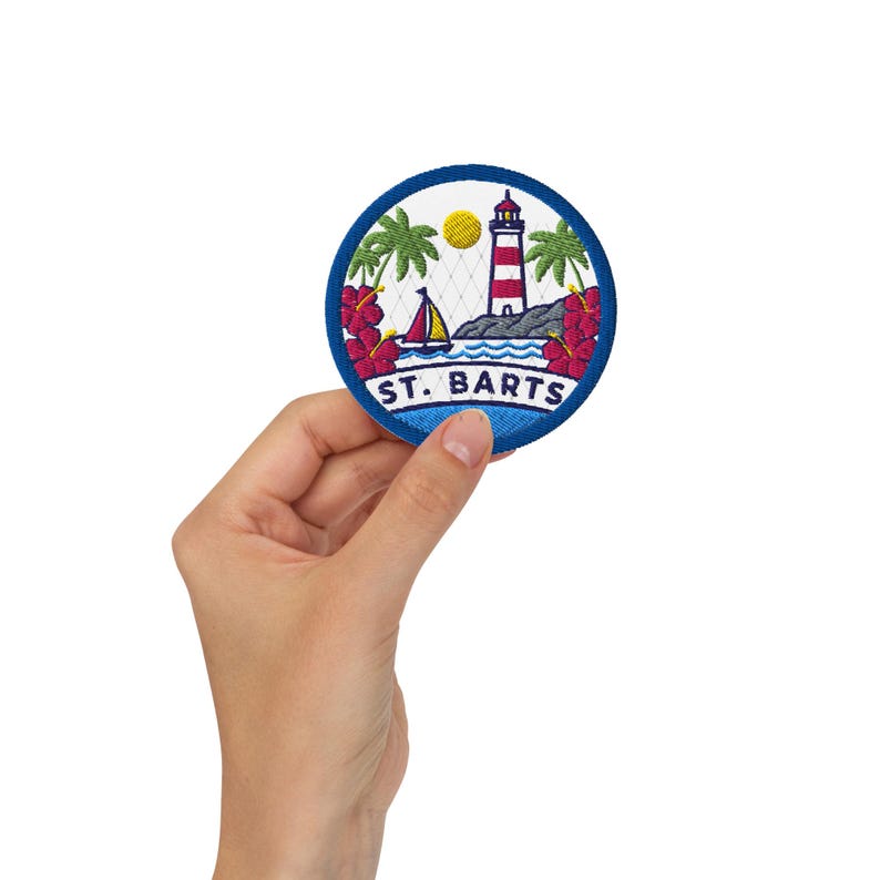 May include: A circular embroidered patch featuring a red and white lighthouse, a sailboat, green palm trees, and red hibiscus flowers against a white background. The text ST. BARTS is embroidered in blue at the bottom within a blue border.