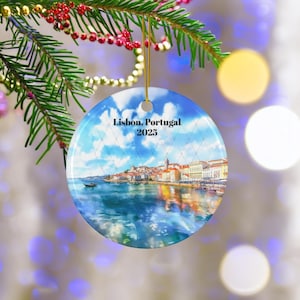 May include: Round Christmas ornament featuring a watercolor image of Lisbon, Portugal, with the text "Lisbon, Portugal 2025" against a blue sky background. The ornament is hanging from a gold string, with a blurred background.