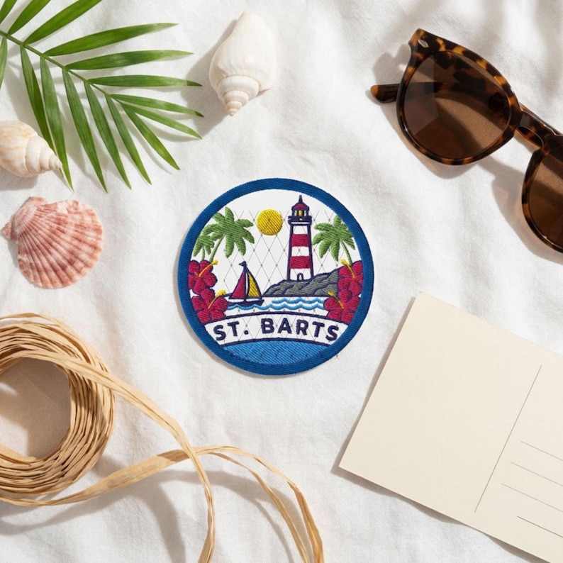 May include: A circular embroidered patch featuring a red and white lighthouse, a sailboat, palm trees, and red hibiscus flowers. The text ST. BARTS is embroidered in blue at the bottom of the patch, which has a dark blue border.