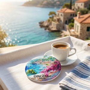Peut inclure: Un dessous de verre rond en c&eacute;ramique illustrant une sc&egrave;ne c&ocirc;ti&egrave;re color&eacute;e avec des voiliers, des fleurs roses et le texte "C&ocirc;te d'Azur 2026" imprim&eacute;, pos&eacute; &agrave; c&ocirc;t&eacute; d'une tasse en c&eacute;ramique blanche remplie de caf&eacute; noir sur un tissu blanc.