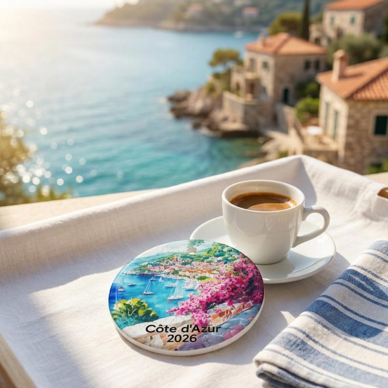 Peut inclure: Un dessous de verre rond en c&eacute;ramique illustrant une sc&egrave;ne c&ocirc;ti&egrave;re color&eacute;e avec des voiliers, des fleurs roses et le texte "C&ocirc;te d'Azur 2026" imprim&eacute;, pos&eacute; &agrave; c&ocirc;t&eacute; d'une tasse en c&eacute;ramique blanche remplie de caf&eacute; noir sur un tissu blanc.