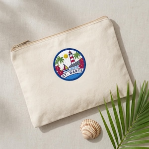 May include: A beige canvas zippered pouch featuring a circular embroidered patch. The patch displays a red and white lighthouse, a sailboat, palm trees, and red hibiscus flowers, with the text ST. BARTS printed in blue at the bottom.