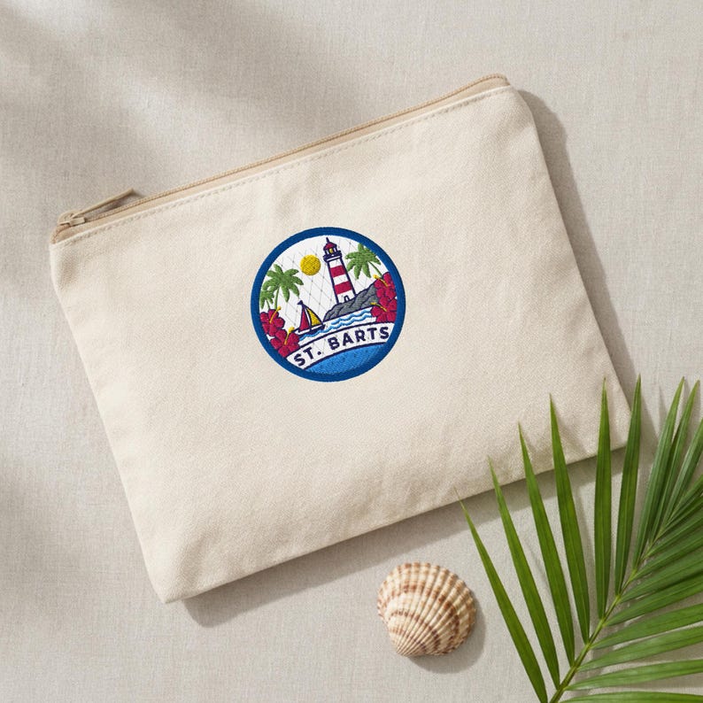 May include: A beige canvas zippered pouch featuring a circular embroidered patch. The patch displays a red and white lighthouse, a sailboat, palm trees, and red hibiscus flowers, with the text ST. BARTS printed in blue at the bottom.