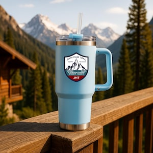 Könnte beinhalten: Ein hellblauer isolierter Becher mit Henkel und transparentem Deckel. Der Becher zeigt ein John Muir Trail-Logo mit Text und Berggrafiken. Der Hintergrund zeigt eine Bergkette und Bäume.