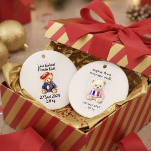 May include: Two white ceramic Christmas ornaments with teddy bear illustrations, each in a gift box with a red bow. One ornament has a bear in a blue jacket and red beret, the other in a pink sweater. Each has a name, date, and weight.
