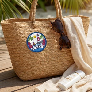 May include: A woven straw tote bag featuring a circular embroidered patch with a lighthouse, sailboat, palm trees, and the text ST. BARTS. The bag is adorned with brown tortoiseshell sunglasses and a white fabric cloth draped over the side.
