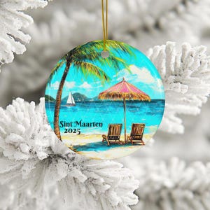 May include: A round Christmas ornament with a beach scene. The ornament features a palm tree, a beach umbrella, two lounge chairs, and a sailboat in the distance. The text "Sint Maarten 2025" is printed on the ornament.