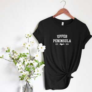 May include: Black t-shirt with white text that reads "Upper Peninsula" with a white outline of the Upper Peninsula of Michigan and the text "Est. 1819".