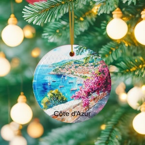Peut inclure: Une d&eacute;coration de No&euml;l ronde en c&eacute;ramique illustrant une peinture &agrave; l'aquarelle de la c&ocirc;te d'Azur avec une mer bleue, des voiliers, un village c&ocirc;tier et des fleurs de bougainvillier rose vif. Le texte C&ocirc;te d'Azur est inscrit en bas.