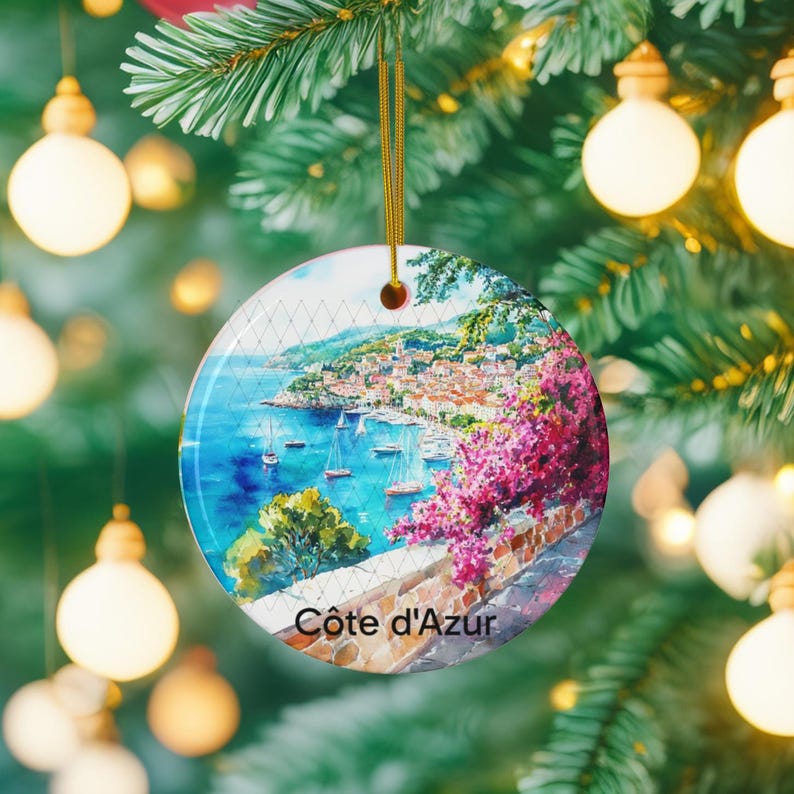 Peut inclure: Une d&eacute;coration de No&euml;l ronde en c&eacute;ramique illustrant une peinture &agrave; l'aquarelle de la c&ocirc;te d'Azur avec une mer bleue, des voiliers, un village c&ocirc;tier et des fleurs de bougainvillier rose vif. Le texte C&ocirc;te d'Azur est inscrit en bas.