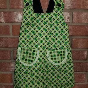 May include: A green and black plaid apron with shamrock patterns and two pockets trimmed with white lace. The apron has black straps and buttons, and is hanging against a brick wall.