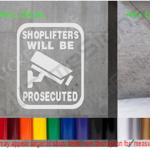 May include: White vinyl decal with the text "Shoplifters Will Be Prosecuted" and a security camera icon. The decal is on a gray concrete wall.