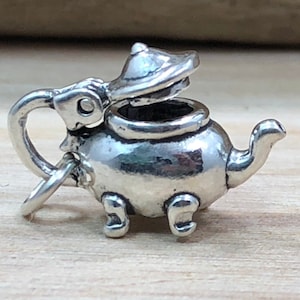 May include: A small, silver-colored teapot charm with an open lid. The teapot has a handle, a spout, and sits on three small feet. The charm is likely made of metal and is designed to be attached to a bracelet or necklace.