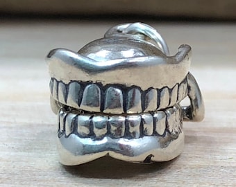 May include: A silver-toned charm in the shape of a set of dentures. The charm has detailed teeth and a rounded top, with a small loop for attaching to a bracelet or necklace. The charm is made of a shiny metal.