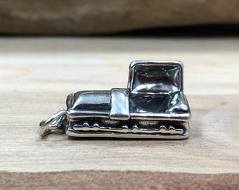 May include: A small, silver-colored charm in the shape of a miniature coffin. The charm has a hinged lid and a detailed design, including a clasp and decorative elements. The charm is attached to a small loop for use as a pendant or charm.