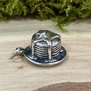 May include: A silver-toned charm in the shape of a stack of pancakes with syrup dripping down. The charm sits on a small plate and has a loop for attaching to a necklace or bracelet. The background is a wooden surface.