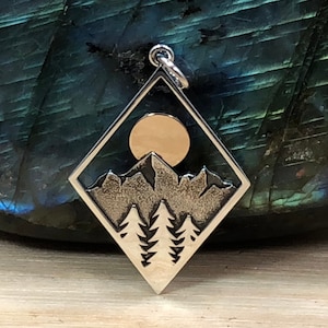 May include: A silver pendant with a mountain and forest scene inside a diamond-shaped frame. A gold-colored circle represents the sun or moon. The pendant has a bail for a chain.