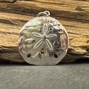 May include: A polished silver-toned pendant shaped like a sand dollar, featuring a central five-pointed star pattern and five small circular perforations. The pendant includes a small circular jump ring at the top for attachment to a necklace chain.
