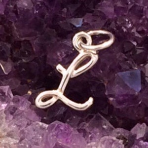 May include: A silver-colored cursive letter "L" charm, with a loop for attaching to a necklace or bracelet. The charm is displayed against a background of a purple crystal formation. The text "Millpond Jewelry Co" is in the bottom right corner.