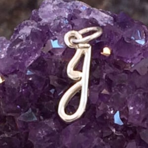 May include: A silver-colored pendant in the shape of the letter "J" hangs against a backdrop of a deep purple amethyst crystal. The pendant is a delicate, cursive design. The text "Millpond Jewelry Co" is visible at the bottom.