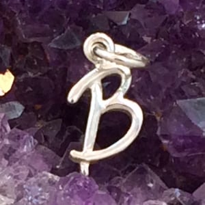 May include: A silver-colored charm in the shape of the letter "B" hangs against a backdrop of a deep purple amethyst crystal. The charm is a delicate, cursive font, and the amethyst has a crystalline structure.