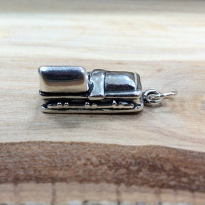 May include: A small, silver-colored charm in the shape of a miniature suitcase. The charm has a rectangular body with a curved top and detailed edges. A small loop is attached for hanging. The charm is set against a light brown wooden surface.