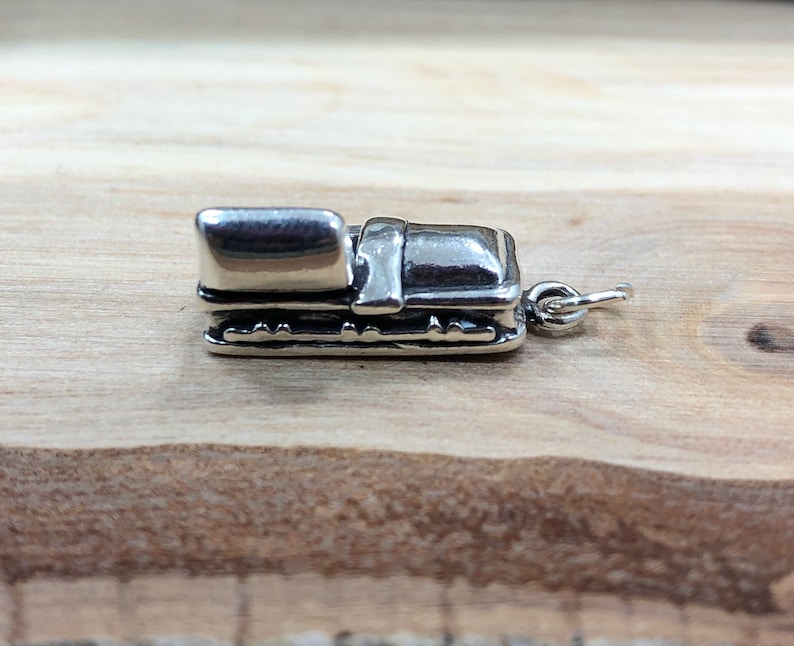 May include: A small, silver-colored charm in the shape of a miniature suitcase. The charm has a rectangular body with a curved top and detailed edges. A small loop is attached for hanging. The charm is set against a light brown wooden surface.