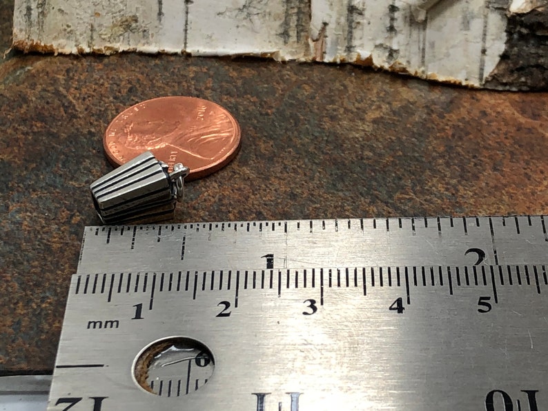 Może przedstawiać: Srebrny charm w kształcie wiaderka popcornu, o długości około 2,5 cm, pokazany obok linijki i pensa, aby pokazać skalę.