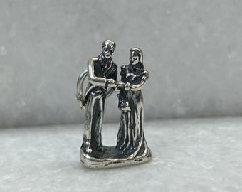 May include: A silver charm depicting a bride and groom standing together. The groom is wearing a suit and the bride is wearing a dress. The charm is a wedding-themed decoration.