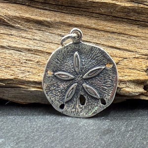 May include: A circular silver pendant shaped like a sand dollar, featuring a textured surface with a central five-petaled star pattern and small perforations along the edges. The pendant includes a small silver jump ring at the top for attachment.