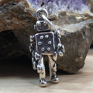 May include: A silver charm in the shape of a robot. The robot has a round head with a face and a body with rivets. The charm is standing on a wooden surface.