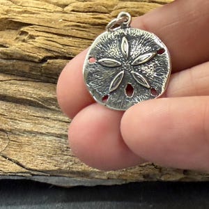 May include: A circular silver-toned pendant featuring a textured surface and a raised five-petal floral design. The pendant has a small jump ring attached to the top edge, allowing it to be suspended from a chain or cord for use as jewelry.