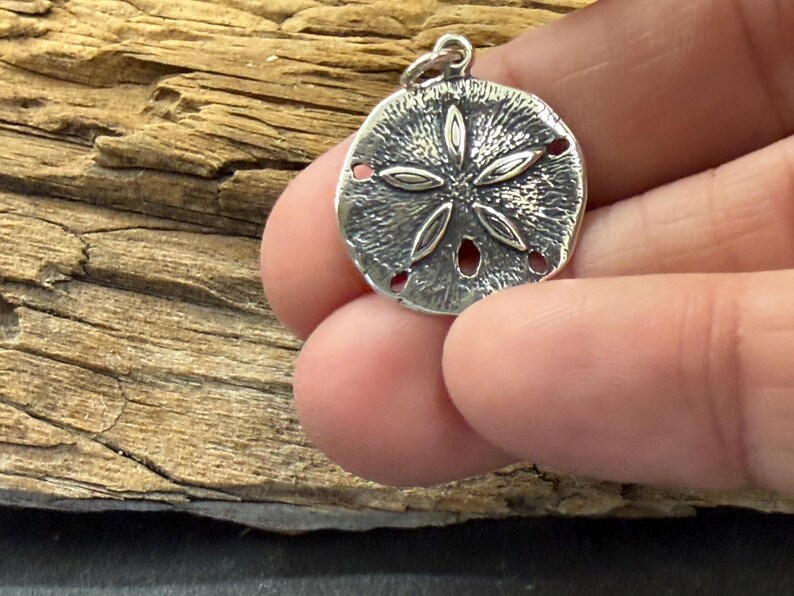 May include: A circular silver-toned pendant featuring a textured surface and a raised five-petal floral design. The pendant has a small jump ring attached to the top edge, allowing it to be suspended from a chain or cord for use as jewelry.