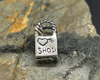 May include: A small, silver-colored charm in the shape of a shopping bag. The charm has a twisted handle and the words "I ♡ to SHOP" are engraved on the front. The charm is set against a textured gray stone background.
