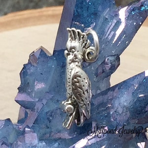 May include: A silver-colored charm in the shape of a cockatoo, perched on a branch. The bird has detailed feather and facial features. The charm is set against a backdrop of blue crystals and a wooden surface.